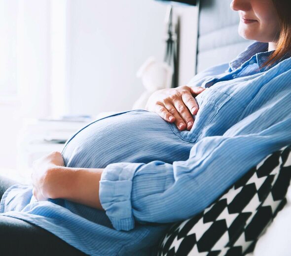 Kobieta w ciąży siedzi na łóżku i opiera się o zagłówek i biało czarną poduszkę. Ma blond włosy i niebieską tunikę. Rękami obejmuje brzuch. 