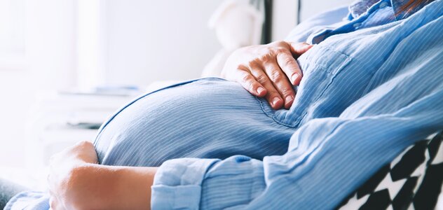 Kobieta w ciąży siedzi na łóżku i opiera się o zagłówek i biało czarną poduszkę. Ma blond włosy i niebieską tunikę. Rękami obejmuje brzuch. 