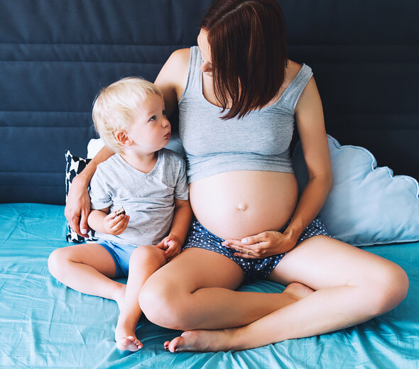 Mama w ciąży siedzi z synkiem na łóżku na niebieskim prześcieradle i opiera się o zagłówek. Patrzy na synka. Obejmuje go ręką. Ma szarą podwiniętą koszulkę na ramiączka i granatowe krótkie spodenki w białe groszki.  Synek ma blond włosy, szarą koszulkę i niebieskie krótkie spodenki. 