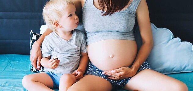 Mama w ciąży siedzi z synkiem na łóżku na niebieskim prześcieradle i opiera się o zagłówek. Patrzy na synka. Obejmuje go ręką. Ma szarą podwiniętą koszulkę na ramiączka i granatowe krótkie spodenki w białe groszki.  Synek ma blond włosy, szarą koszulkę i niebieskie krótkie spodenki. 