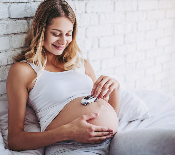 Kobieta w ciąży siedzi na łóżku na białej pościeli i opiera się o białą ścianę. Ma na sobie biały stanik i szare spodnie. Ma blond długie włosy. Uśmiecha się. Trzyma mały biały samochodzik na brzuchu. 