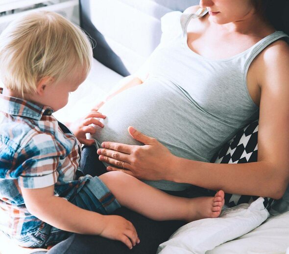 Kobieta w ciąży siedzi na łóżku i opiera się o zagłówek. Dotyka rękami brzucha. Ma wyprostowane nogi. Na jej nogach siedzi mały chłopczyk. Kobieta ma szarą koszulkę na ramiączkach . Chłopiec ma blond włoski, koszulę w kratkę z krótkim rękawkiem i niebieskie dżinsy. 