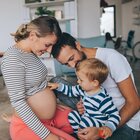 Mama, tata i synek siedzą na podłodze. Śmieją się. Chłopiec podciąga bluzkę mamy i ogląda jej ciążowy brzuszek. Chłopiec ubrany jest w śpioszki w paski, tata ma białą koszulkę a mama ma bluzkę w biało-czarne paski. W tle widać nowoczesne minimalistyczne wnętrze i rower. 