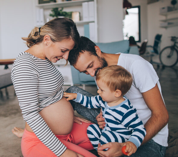 Mama, tata i synek siedzą na podłodze. Śmieją się. Chłopiec podciąga bluzkę mamy i ogląda jej ciążowy brzuszek. Chłopiec ubrany jest w śpioszki w paski, tata ma białą koszulkę a mama ma bluzkę w biało-czarne paski. W tle widać nowoczesne minimalistyczne wnętrze i rower. 