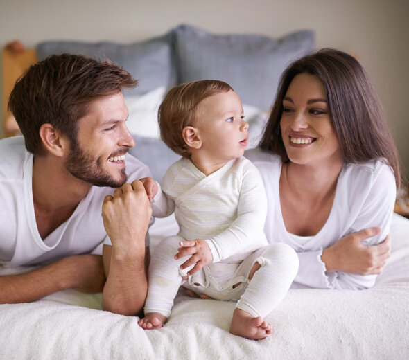 Mama i tata leżą na łóżku na brzuchu. Pomiędzy nimi siedzi niemowlak. Tata trzyma dziecko za rączkę. Rodzice śmieją się. Wszyscy ubrani są na biało. 