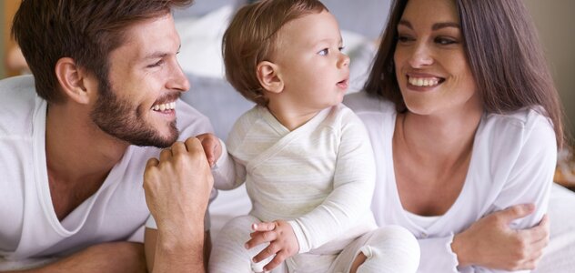 Mama i tata leżą na łóżku na brzuchu. Pomiędzy nimi siedzi niemowlak. Tata trzyma dziecko za rączkę. Rodzice śmieją się. Wszyscy ubrani są na biało. 