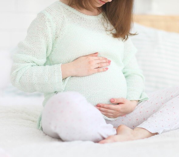 Na łóżku siedzi ze skrzyżowanymi nogami młoda kobieta w ciąży. Trzyma się za brzuszek. Na sobie ma jasno zielony sweter i spodnie od piżamy w różowe kropki. 