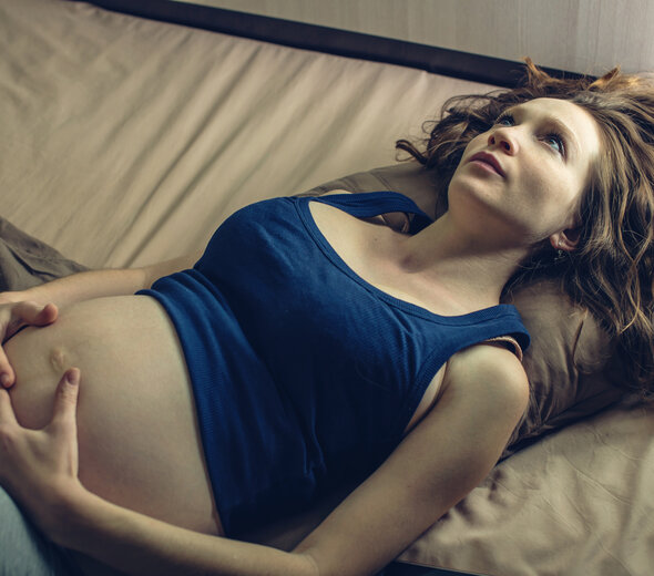 Młoda kobieta w ciąży leży na łóżku na plecach i przygnębiona patrzy w sufit. Rękami trzyma swój brzuszek. Ma na sobie granatowy top i spodnie od dresu. 
