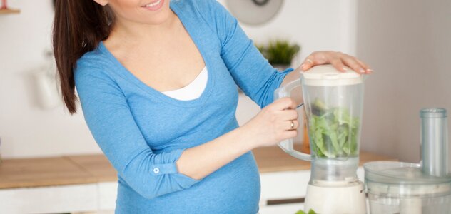 Młoda kobieta w ciąży stoi w kuchni. Przygotowuje sobie smoothie z zielonych liści w blenderze. Na stole leżą przygotowane i pokrojone warzywa i owoce. Kobieta ma sobie niebieska bluzkę. . 