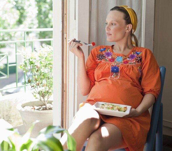 Młoda kobieta w ciąży siedzi na krześle przy oknie i je sałatkę. Talerz trzyma na kolanach. Jest ubrana w pomarańczową sukienkę, na głowie ma żółtą opaskę. 
