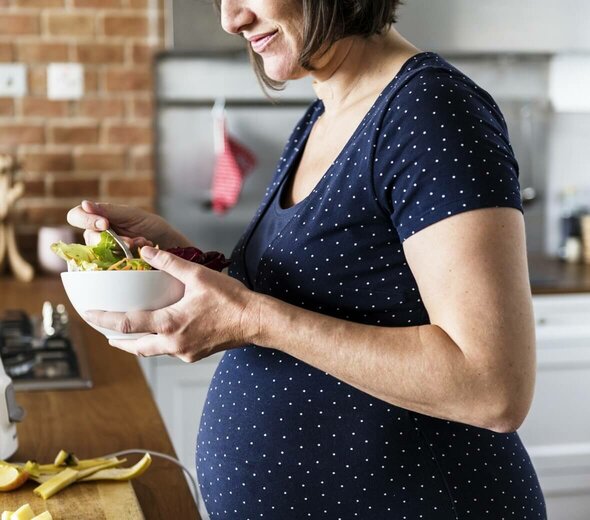 Kobieta w ciąży stoi w kuchni i je sałatkę z porcelanowej miseczki. Jest ubrana w granatową bluzkę w białe kropki. W tle widać ceglaną ścianę. 