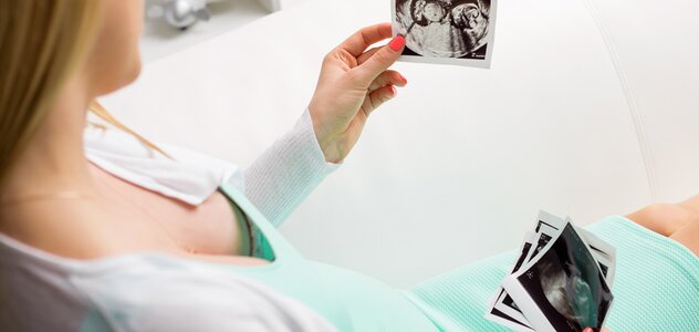 Kobieta w ciąży siedzi z wyciągniętymi nogami na kanapie i ogląda zdjęcia USG. Ma na sobie miętową sukienkę i biały, rozpinany sweterek. 