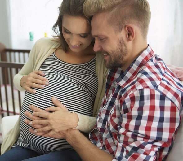 Młoda para siedzi na kanapie. Kobieta jest w zaawansowanej ciąży. Oboje patrzą na jej brzuszek, dotykają go i uśmiechają się szczęśliwi. W tel widać dziecięce łóżeczko ze szczebelkami. 