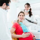 Kobieta w ciąży siedzi na fotelu dentystycznym. Asystentka lekarza zakłada jej śliniak jednorazowy. Dentysta ma na sobie maseczkę i okulary. Rozmawiają i uśmiechają się do siebie. 