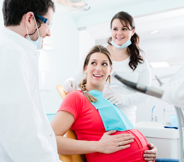 Kobieta w ciąży siedzi na fotelu dentystycznym. Asystentka lekarza zakłada jej śliniak jednorazowy. Dentysta ma na sobie maseczkę i okulary. Rozmawiają i uśmiechają się do siebie. 