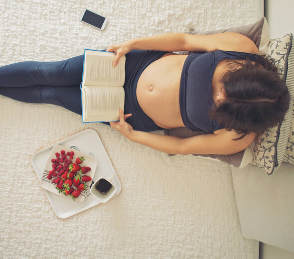 Młoda kobieta w ciąży leży na łóżku i czyta książkę. Ma na sobie granatowy top i granatowe legginsy. Obok niej leżą telefon i owoce na talerzu. 