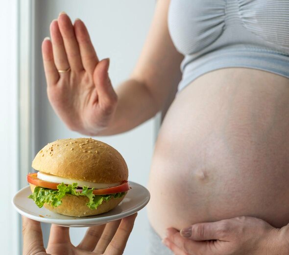 Kobieta w ciąży stanowczym gestem dłoni odmawia zjedzenia bułki. Bułka leży na talerzyku, w środku jest ser, pomidor i sałata.  Kobieta ma szary biustonosz sportowy i nagi brzuch. 