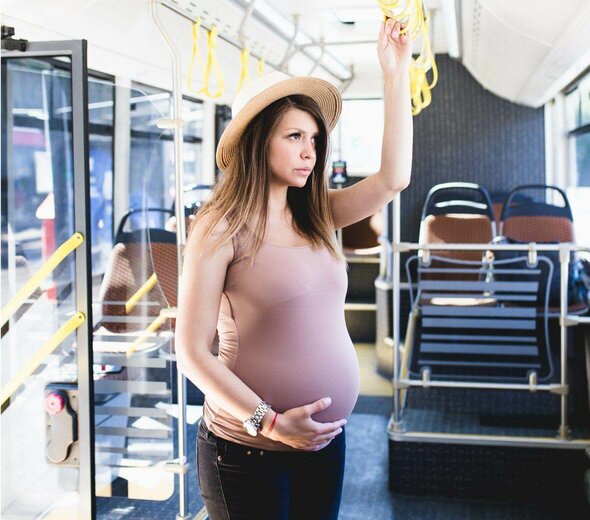 Kobieta w ciąży stoi na środku pustego autobusu i trzyma się poręczy. Drugą dłonią podtrzymuje brzuszek. Jest zamyślona, patrzy przez okno. Na głowie ma letni kapelusz. 