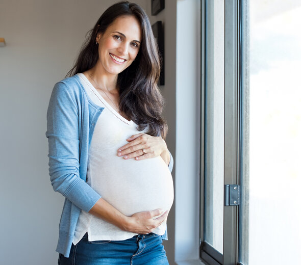 Kobieta w zaawansowanej ciąży uśmiecha się do obiektywu, obejmując z czułością swój brzuszek. Ma długie, ciemne włosy, na sobie białą bluzkę i niebieski rozpinany sweterek. 