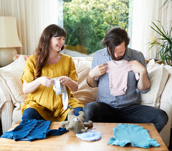 Kobieta w ciąży wraz z przyszłym tatą siedzą na kanapie i oglądają ubranka dla dziecka. Ojciec trzyma różowe body na krótki rękawek jakby chciał je przymierzyć.
