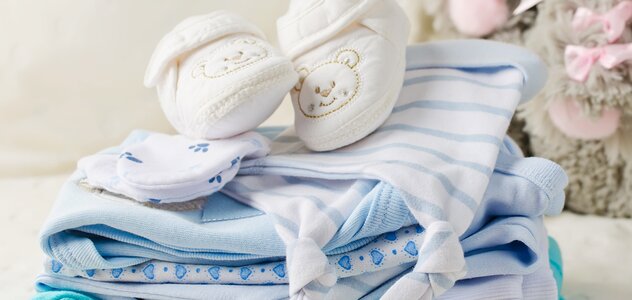 Na łóżku leży stosik ubranek przygotowanych dla przyszłego dziecka, między innymi: niebieskie bluzeczki, białą czapeczkę, rękawiczki oraz buciki z misiami.