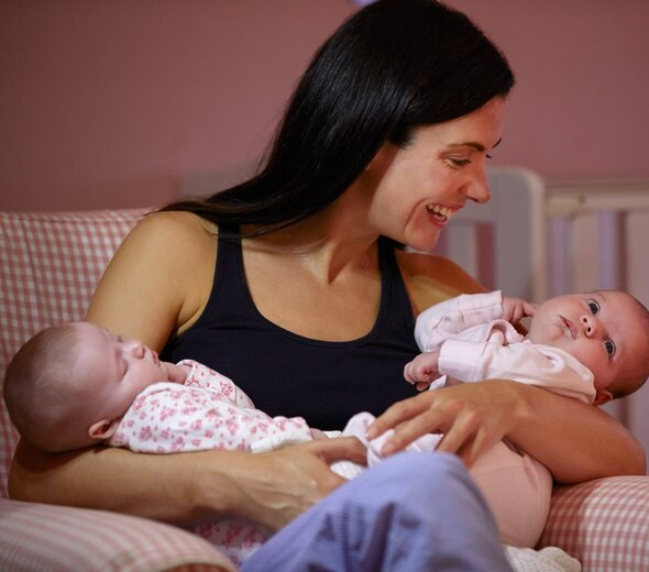 Matka siedzi w fotelu w kratę trzymając jednocześnie dwójkę niemowląt. Jedno z dzieci śpi, a drugie rozgląda się. Matka uśmiecha się szczerze i przytula je do siebie.