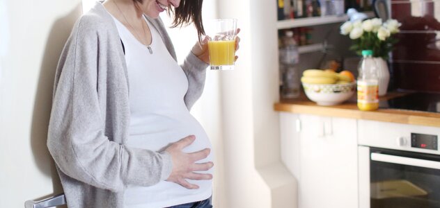 Kobieta w ciąży stoi w kuchni oparta o lodówkę. W jednej ręce trzyma szklankę z sokiem pomarańczowym, a drugą ma położoną na brzuchu. Przyszłą mama uśmiecha się.