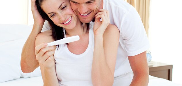 Mężczyzna przytula od tyłu kobietę, która trzyma w ręku test ciążowy. Oboje uśmiechają się. Para wygląda na szczęśliwą z faktu, że ich rodzina się powiększy.