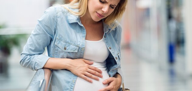 Kobieta w ciąży łapie się rękoma za brzuch z powodu bólu i ma bolesny wyraz twarzy. Jest ubrana w białą podkoszulkę, którą częściowo zakrywa niebieska koszula. 