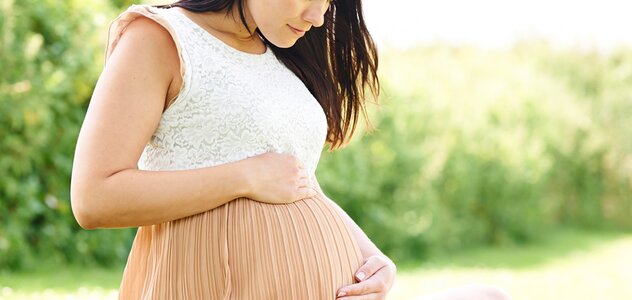 Przyszła mama, ubrana w jasną sukienkę, siedzi na trawie trzymając się czule za brzuch. Prowadzenie zdrowego stylu życia jest bardzo ważne również w czasie ciąży.