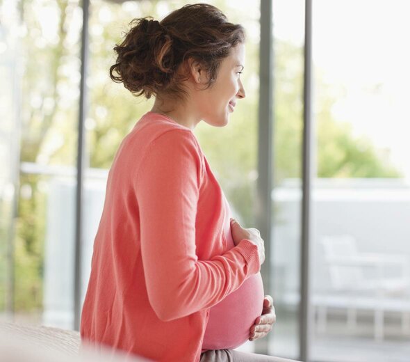 Przyszła mama stoi zamyślona na balkonie i uśmiecha się. Młoda kobieta trzyma się czule za brzuch. Ubrana jest w różową koszulkę, różowy sweter i luźne, szare spodnie.