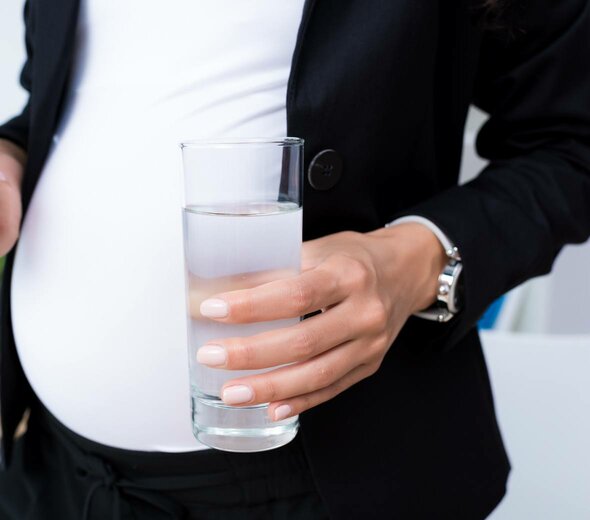 Kobieta w ciąży stoi ze szklanką w ręku. Przyszła mama ubrana jest w czarne, wygodne spodnie, białą koszulkę i czarną marynarkę, a jej nadgarstek zdobi srebrny zegarek.