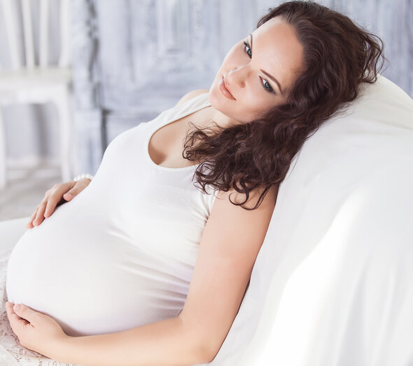 Przyszła mama siedzi wygodnie oparta na kanapie. Kobieta trzyma się czule za brzuch. Odpowiednia ilość wypoczynku w czasie ciąży minimalizuje ryzyko komplikacji.
