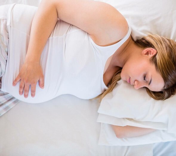 Przyszła mama śpi na łóżku, trzymając się jedną ręką za brzuch. Kobieta jest ubrana na biało i wygląda na spokojną. Odpowiednia ilość snu jest ważna w czasie ciąży.