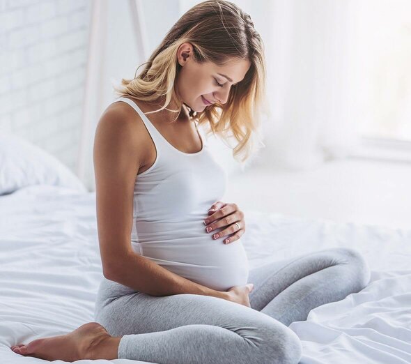 Przyszła mama siedzi wygodnie na łóżku ubrana w szare, dresowe spodnie oraz białą koszulkę bez rękawów. Kobieta czule obejmuje brzuch i uśmiecha się patrząc na niego.