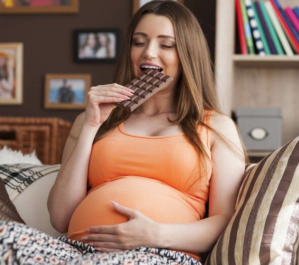 Młoda kobieta w ciąży siedzi wygodnie na łóżku w pomarańczowej koszulce bez rękawów i kolorowych spodniach w kwiaty. Przyszła mama trzyma w ręku tabliczkę czekolady.