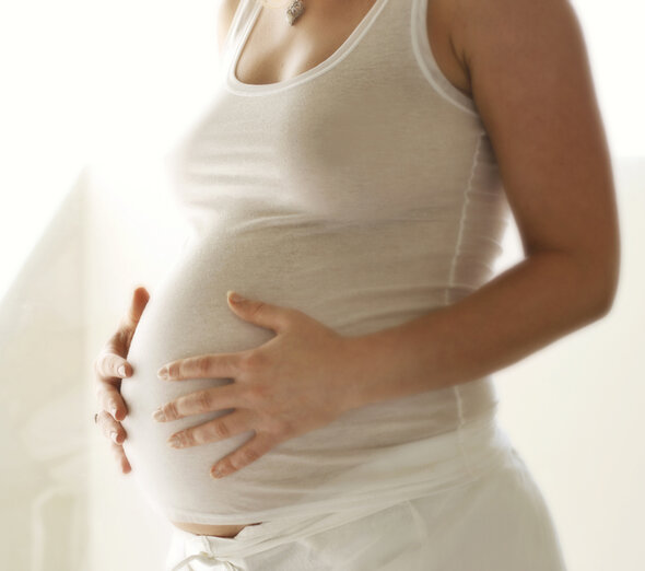 Młoda kobieta w ciąży wygodnie stoi ubrana w białe, luźne spodnie oraz jasną koszulkę bez rękawów. Przyszła mama delikatnie i czule trzyma się za swój brzuch.