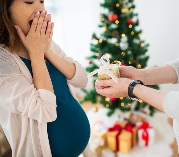 Kobieta w ciąży stoi na przeciwko mężczyzny. Ma brązowe, proste, półdługie włosy, granatową bluzkę i biały rozpinany sweter. Zasłania usta dłońmi i się śmieje. Jest mile zaskoczona. Mężczyzna trzyma w rękach niewielki prezent. W tle widać choinkę. 