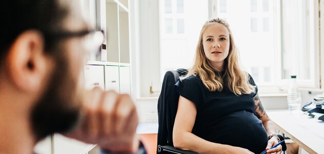 Kobieta w ciąży siedzi na krześle przy biurku w biurze, na tle okna. Patrzy na siedzącego obok mężczyznę. Ma długie blond włosy i granatową sukienkę. Z boku widać biały regał. 