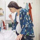 Kobieta w ciąży stoi w łazience nad umywalką i myje zęby. Ma biała bluzkę i szlafrok w kwiaty. Ma ciemne długie upięte włosy. 