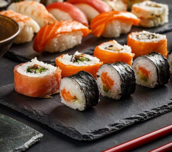 Na czarnej podstawce leżą kawałki sushi, różnego rodzaju danie pokrojone jest na klasyczne krążki. Obok leżą bordowe pałeczki do sushi. 