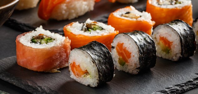 Na czarnej podstawce leżą kawałki sushi, różnego rodzaju danie pokrojone jest na klasyczne krążki. Obok leżą bordowe pałeczki do sushi. 