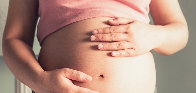 Kobieta w ciąży dotyka dłońmi swój brzuch. Różową bluzkę ma podciągniętą do góry, jej brzuch jest goły. 