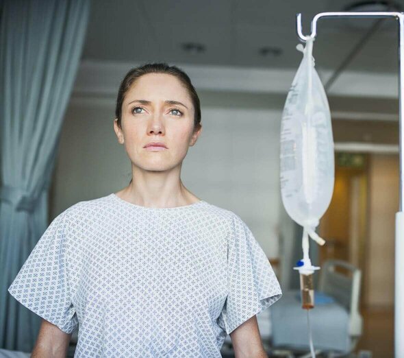 Kobieta stoi w piżamie szpitalnej na korytarzu szpitala. Obok mamy stoi stojak na kroplówkę z podłączonymi witaminami. Kobieta patrzy zatroskana i poważna daleko przed siebie.