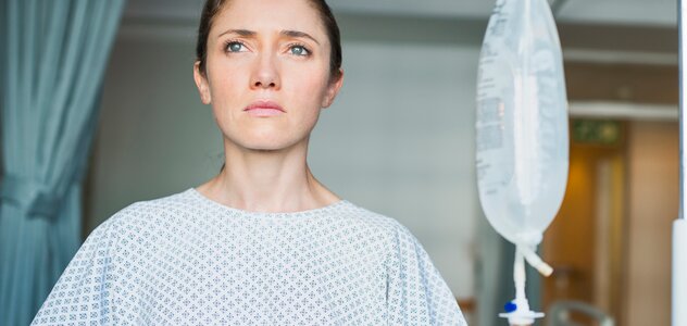 Kobieta stoi w piżamie szpitalnej na korytarzu szpitala. Obok mamy stoi stojak na kroplówkę z podłączonymi witaminami. Kobieta patrzy zatroskana i poważna daleko przed siebie.