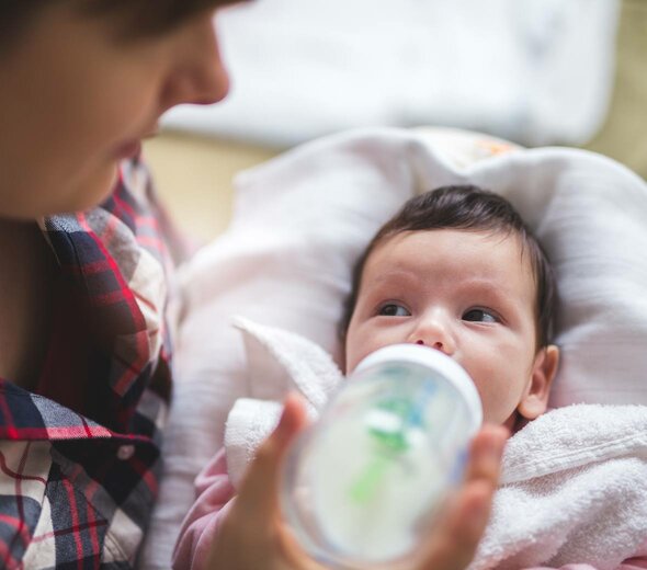 Dziecko jest trzymane przez mamę na rękach i jest karmione butelką. Główka leży na poduszce, a na piersi dziecka jest położony biały ręcznik. Dziecko spogląda na mamę.