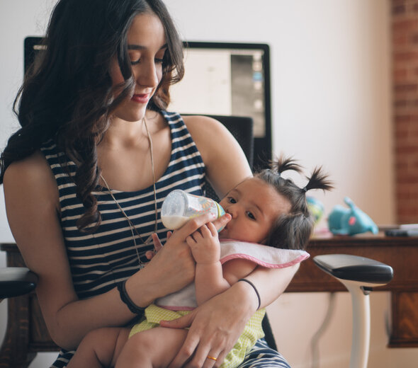 Uśmiechnięta mama siedzi na krześle, trzyma swoją córkę na kolanach i karmi dziecko butelką. Dziewczynka ma założony różowy śliniak i spokojnie patrzy w lewą stronę. 