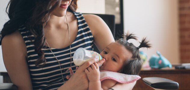 Uśmiechnięta mama siedzi na krześle, trzyma swoją córkę na kolanach i karmi dziecko butelką. Dziewczynka ma założony różowy śliniak i spokojnie patrzy w lewą stronę. 