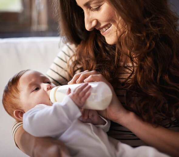 Uśmiechnięta mama trzyma na rękach niemowlę i karmi je butelką. Dziecko ubrane w białe body trzyma butelkę jedną rączką i spokojnie wypija mleko patrząc na butelkę.