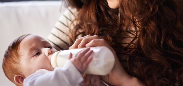 Uśmiechnięta mama trzyma na rękach niemowlę i karmi je butelką. Dziecko ubrane w białe body trzyma butelkę jedną rączką i spokojnie wypija mleko patrząc na butelkę.